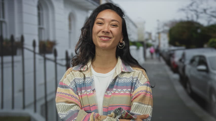 Woman smiling with hands on hips on street in city neighborhood wearing striped jacket and hoop earrings; joyful confidence.