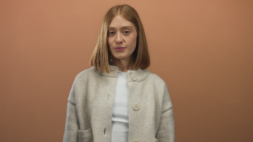 Young blonde woman covers eyes with right hand in bright studio with plain brown wall; playful surprise joy.