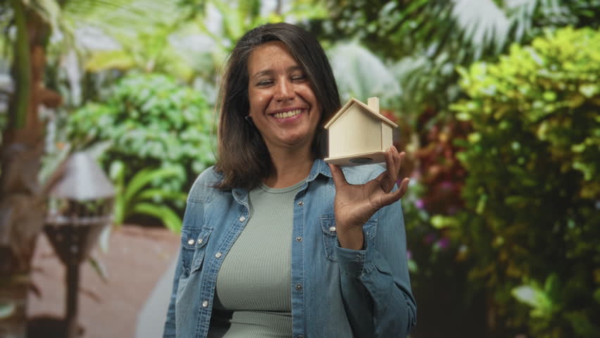 Woman holding wooden house model with hand, smiling in forest setting with denim jacket, plants and lantern visible along pathway; hope.