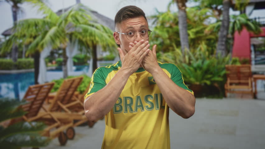 Man wearing yellow brasil soccer shirt points index fingers upward in studio with poolside backdrop; pride fan spirit.