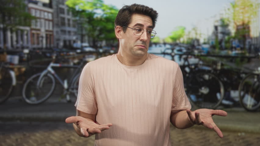 Man in pink t shirt shrugs with palms up by parked bicycles on a canal bridge street, wearing glasses and a neutral expression; bemusement.