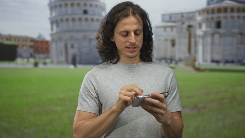 Man holding smartphone taking a selfie in building near the leaning tower of pisa on a grassy plaza; travel curiosity.