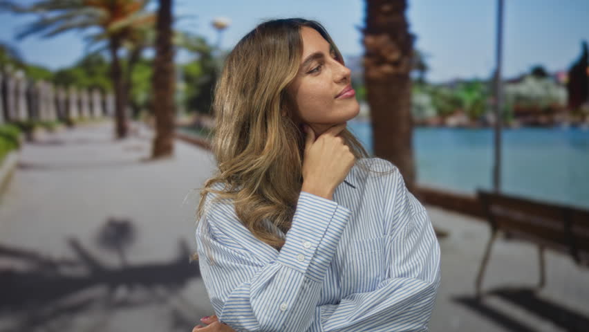 Young blonde woman touches chin with finger on seaside promenade; thoughtfulness confidence introspection.