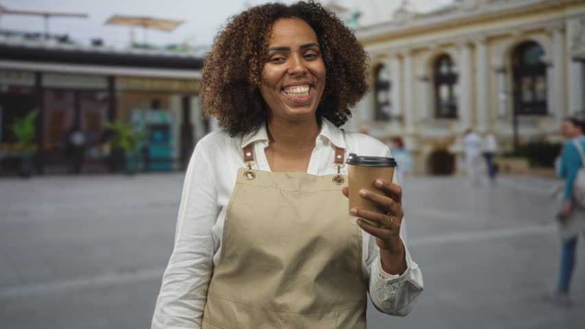 Baker woman holding a coffee cup, lips blowing a kiss in street plaza with historic building; warmth joy.
