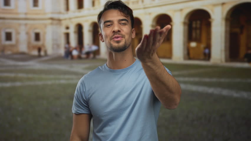 Young hispanic man smiling outdoors at an old university setting, wearing a casual blue shirt, creating a warm and welcoming atmosphere.