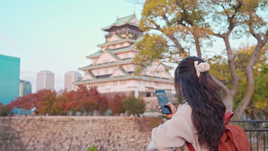 Young female tourist taking a photo of the Osaka Castle, the popular destination in Osaka, Japan