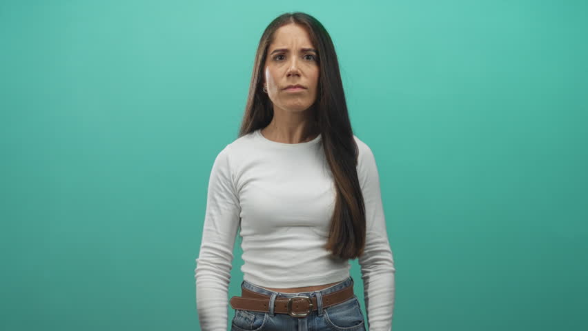Woman cups hands over eyes in studio wearing white top, jeans and belt with long hair visible; playful surprise.