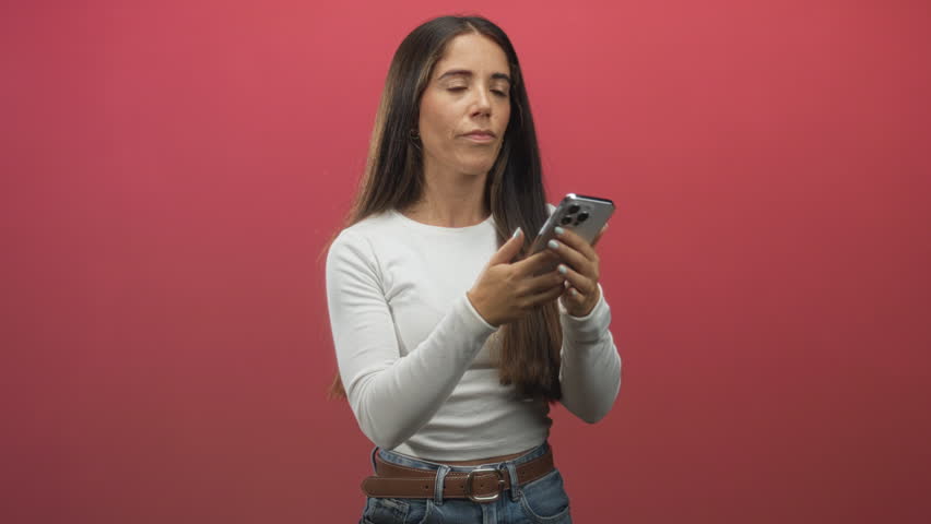Woman holding smartphone to ear, talking and gesturing with hand, wearing white crop top and jeans against red studio backdrop; frustration communication.