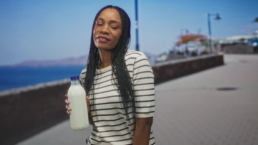 Black woman holding a milk bottle with bare hand on a seaside street promenade, braided hair and striped shirt, slight smile and direct gaze; serenity.