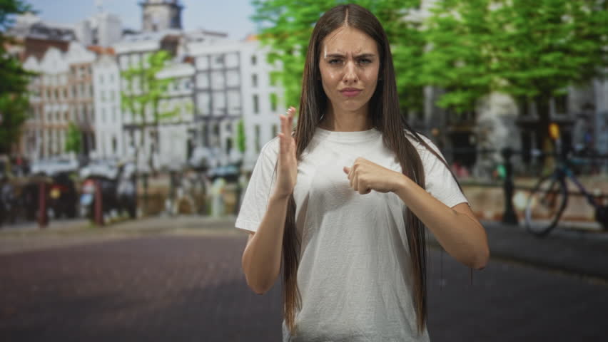 Young woman with long hair showing bare hands making a vertical stop palm and fist tapping gesture on street near canal buildings; defiance boundary.