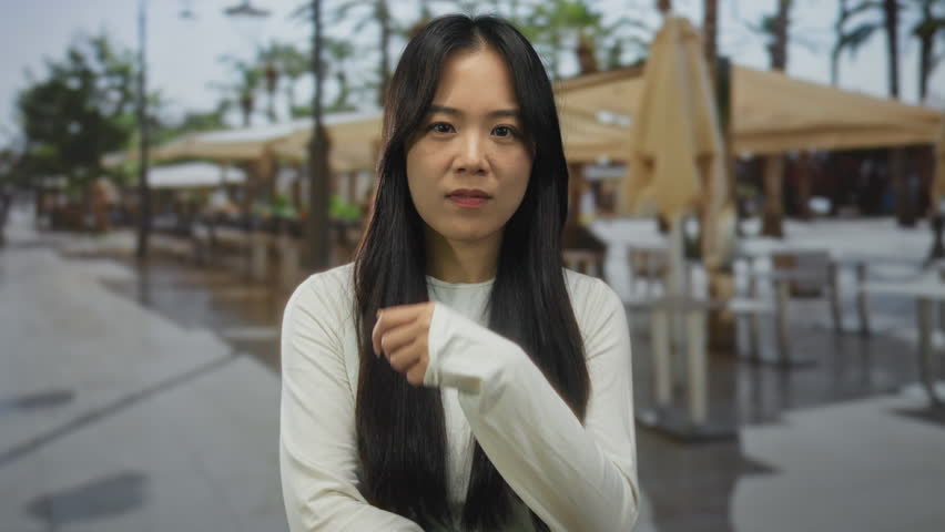 Woman standing outdoors on a restaurant terrace street with a thoughtful expression wearing a white shirt and long black hair surrounded by umbrellas and tables on an urban day.