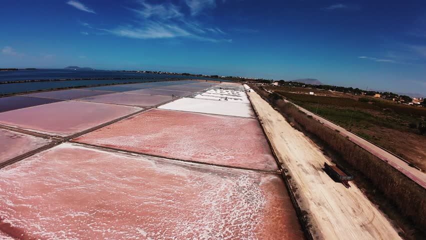 Aerial drone footage of the iconic pink salt evaporation ponds (Saline di Marsala) in the natural reserve near Marsala, Sicily, Italy. The vivid pink hues in the shallow lagoons, created by natural algae and microorganisms, contrast beautifully with turquoise Mediterranean waters, white salt mounds, and traditional windmills. Captured in high-resolution 4K during clear weather, showcasing the abstract patterns and geometric shapes of the salt pans along the coastal landscape. Ideal for travel, nature, abstract, and environmental projects
