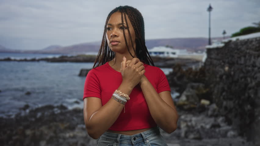 Woman with clasped hands at chest on a seaside street beside a stone wall and lamppost, braided hair visible and bracelets on wrists; serenity.