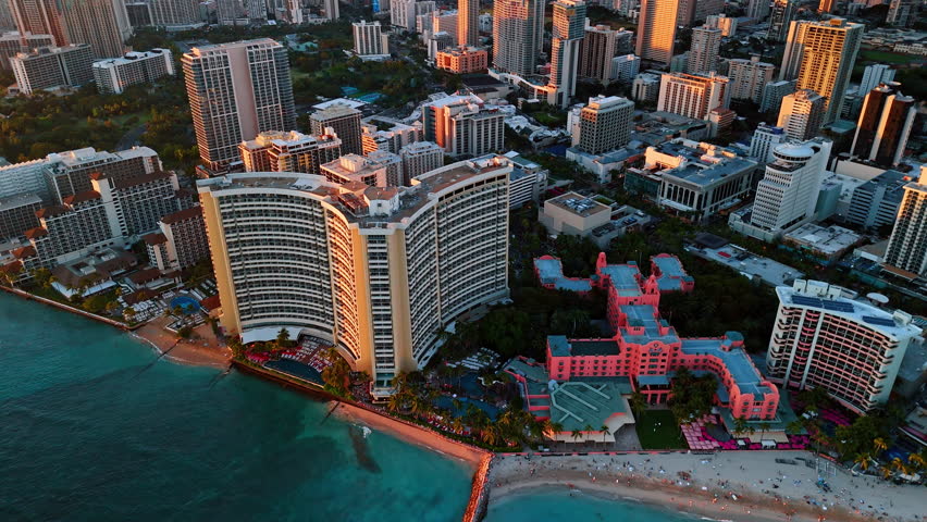 Waves roll to the sandy beach of Waikiki, Honolulu, Hawaii, USA. Beautiful high-rises of the shore are lit with orange light at sunset. Aerial view.