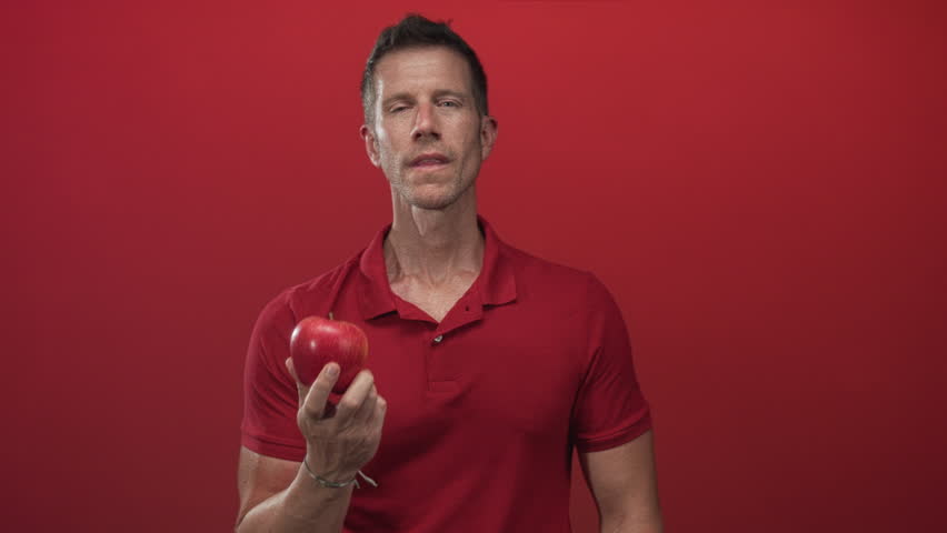Man holding red apple with hand on chest in studio with red backdrop; contemplation temptation sincerity.
