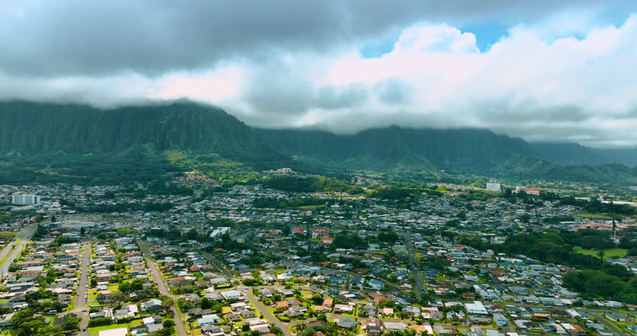 Flying over the cityscape with lush greenery. Ragged verdant mountains are coated with thick clouds at backdrop. Honolulu, Hawaii, USA.