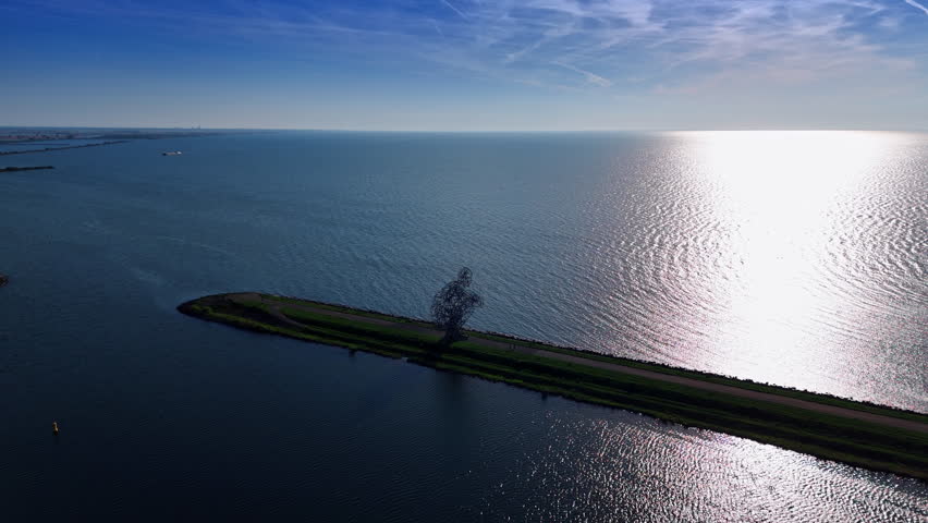 Approaching the metal sculpture of a squatting man on the dam of the Merkemeer Lake. Sun reflects on the surface of the lake. Aerial view. Lelystad, The Netherlands.