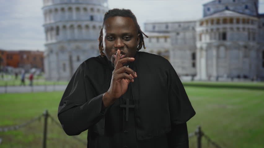 Man in clerical robes wearing a prominent cross necklace, finger to lips for silence in building at the leaning tower of pisa plaza; silence reflection.