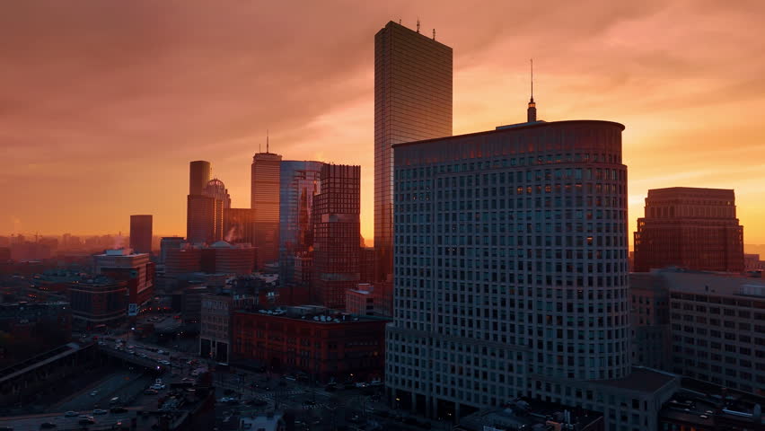 Boston downtown architecture at the backdrop of orange sky. Drone flight over the lively city at sunset.