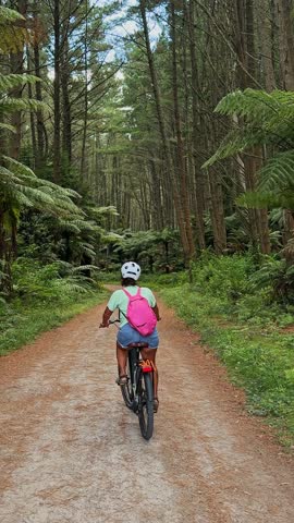 Young black woman cycling on an electric bicycle along a scenic dirt path through a dense pine and fern forest, raising her hand in excitement while exploring the beautiful New Zealand nature