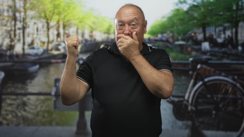 Man covering mouth with hand and raising fist on a canal street beside parked bicycle; surprise contemplation.
