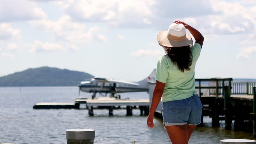 Young woman from behind stretching her arms out in joy on a wooden pier, appreciating the serene lake view with a seaplane and an island in the background during a sunny summer day