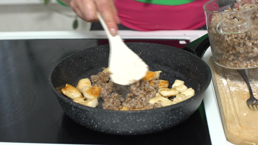 In a kitchen, a person stirs cooked meat and grains in a skillet while another pot holds additional grains ready to add.