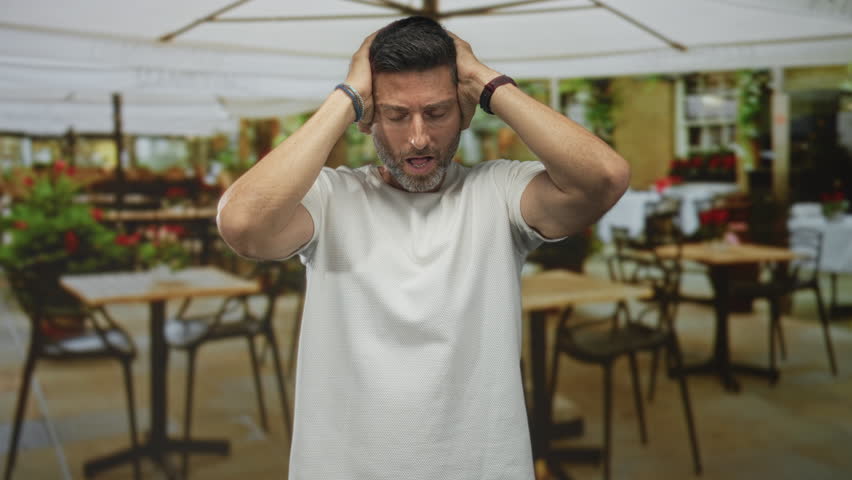 Man holding hands to head covering ears at a street cafe terrace outdoors wearing white t shirt and bracelets; stress and frustration.