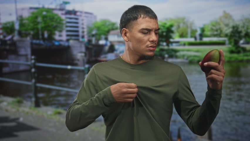 Man holding a mango in one hand with closed eyes and raised gesture by a street waterfront canal, wearing green shirt; contemplation reflection.