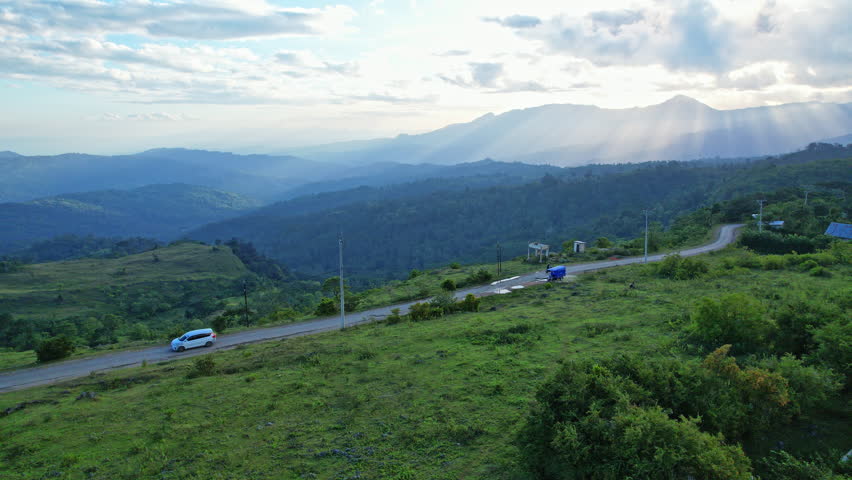 A main mountain road runs through the middle of Timor Island, Indonesia, seen in aerial drone footage as it cuts through dense green forest with dark mountain ridges in the background and sun rays breaking through the clouds over the lush landscape.
