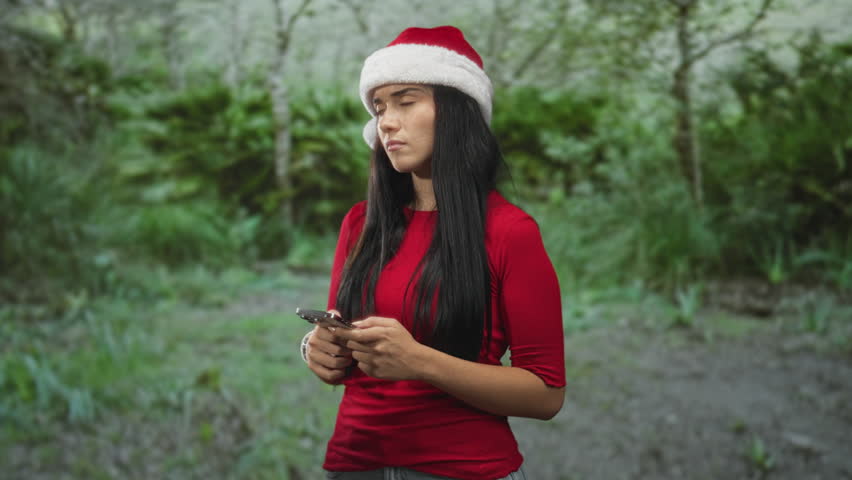 Young woman in a santa hat holding a smartphone with both hands and gazing anxiously upward in a forest; pensive solitude.