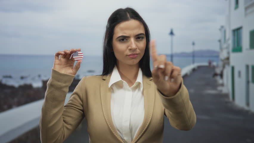 Woman on seaside promenade holding american badge signaling no with finger suggesting refusal or disagreement outdoors
