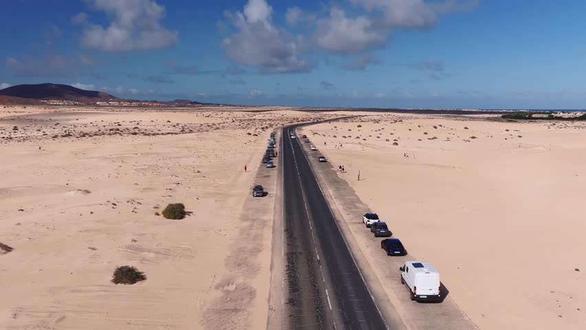 Aerial view shows a two lane road through pale dunes, parked cars, pedestrians, white hotels, Atlantic coast, volcanic hills, bright daylight, cumulus clouds.