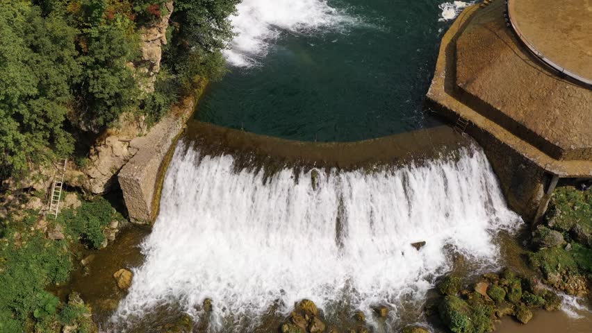 Aerial top-down drone view of a waterfall spilling over a curved stone barrier in Bosnia and Herzegovina. The scene shows white water, textured stone and a calm pool above the drop.