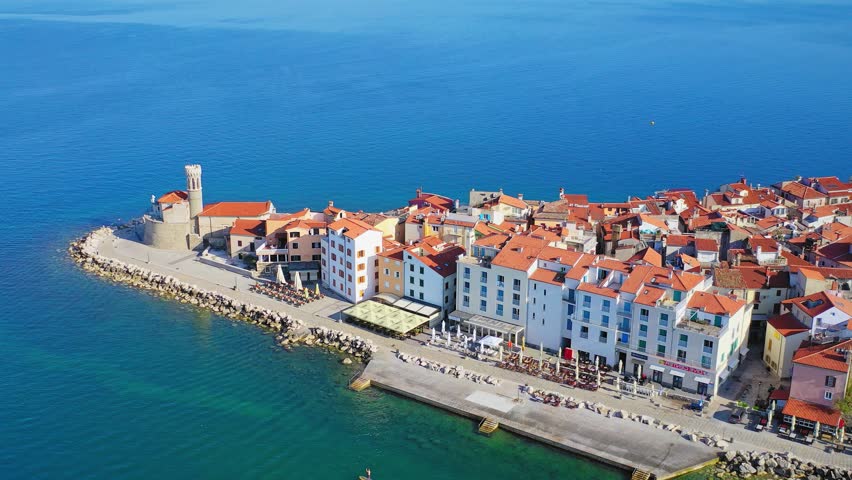Aerial drone view of the colorful waterfront and old town of Piran in Slovenia along the Adriatic coast. The scene shows dense historic buildings, promenade and clear blue sea in a Mediterranean coastal setting.