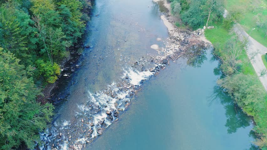 Aerial drone view of a fast-flowing section of a river with rapids and a rocky riverbed in the Kostel area of Slovenia. The scene shows white water, stones and forested banks in a pristine natural environment.