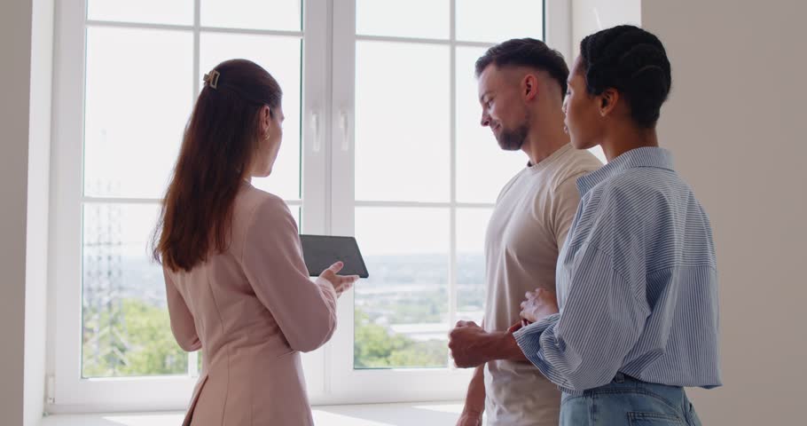 Real estate agent showing tablet screen with contract information to young couple on home tour. People standing by window, professional realtor talking to man and woman to explain details of deal