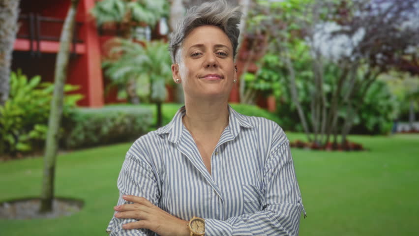 Woman smiling in striped shirt beckons with hand beside red building in lush green park; friendly invitation.