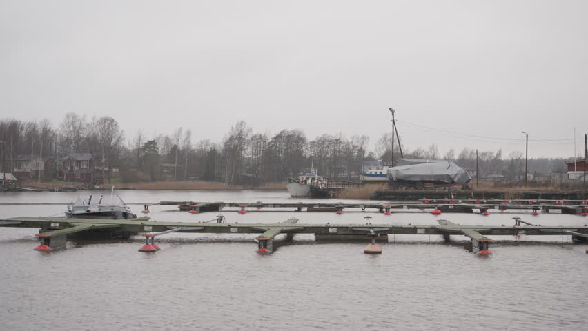 Empty wooden piers and floating docks with red buoys in Hamina marina Finland. Coastal landscape with small motorboat and covered ship on shore under overcast grey sky in autumn season.