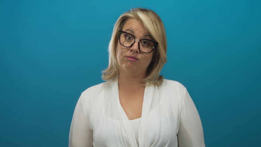 Woman with blonde hair and glasses wearing white blouse pointing fingers to cheeks in studio; playful amusement.
