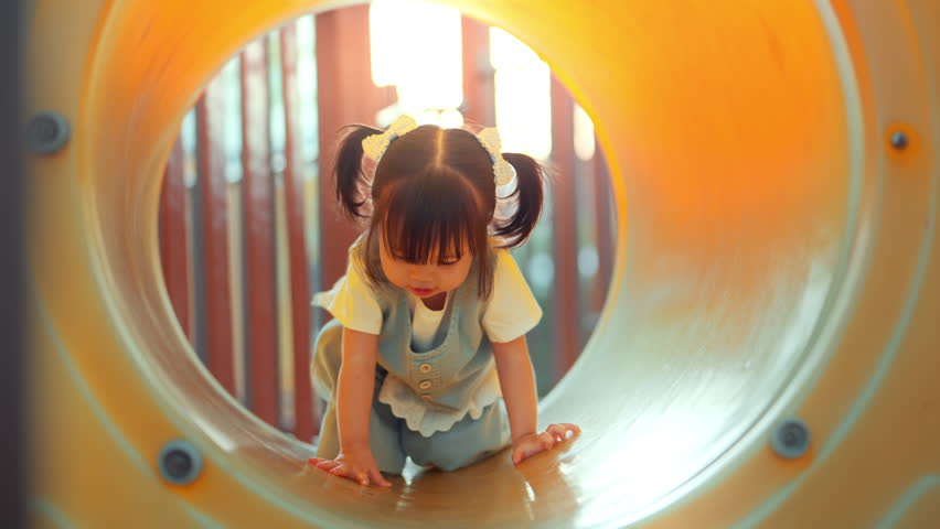Happy cute Asian toddler girl playing and crawling through a yellow tunnel slide at a sunny summer outdoor playground