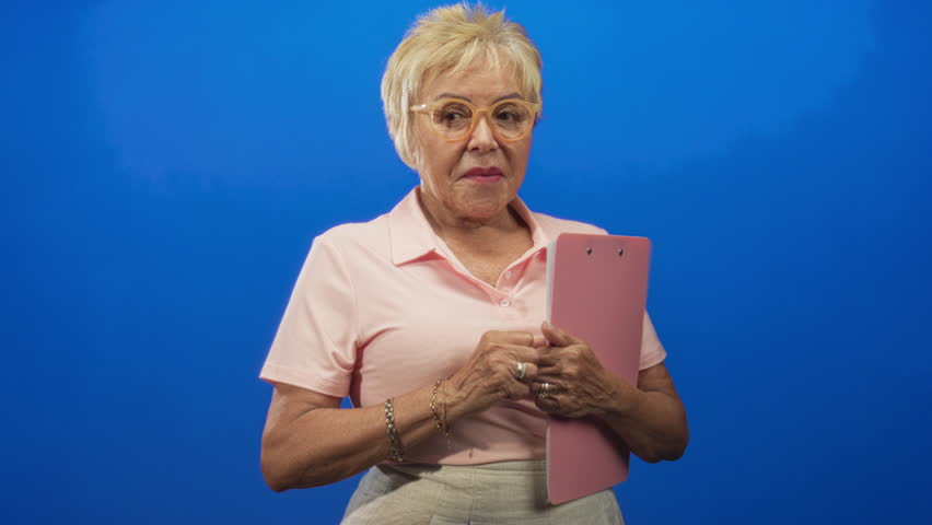 Senior woman wearing pale pink polo and clear glasses holds pink clipboard and clasps hands to chest in studio; playful confidence.