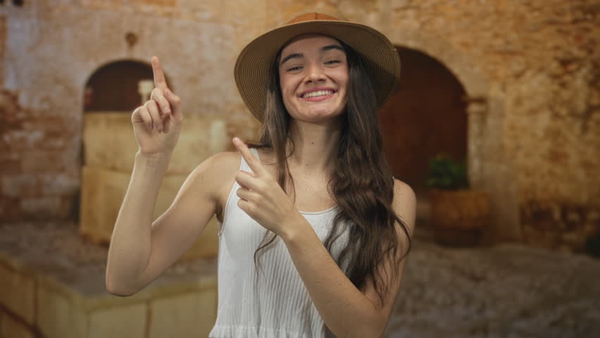 Teenage woman wearing straw hat and white tank top points finger to sunlit street in old town; adventure joy.