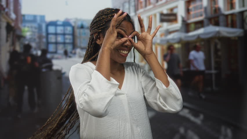 Woman with hand binoculars over eyes wearing a white blouse and braids smiling on a busy street in daylight; playful joy.
