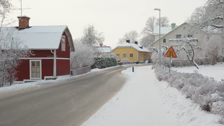 A peaceful winter scene features snow-covered houses lining a winding road, with a car driving through the serene landscape. The vibrant colors of the homes contrast beautifully against the white snow