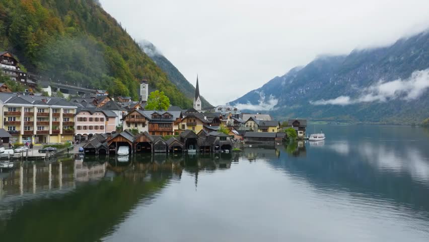 Tranquil Hallstatt village in Austria with colorful houses, church steeple, alpine mountains and boats ferrying tourists across the lake.