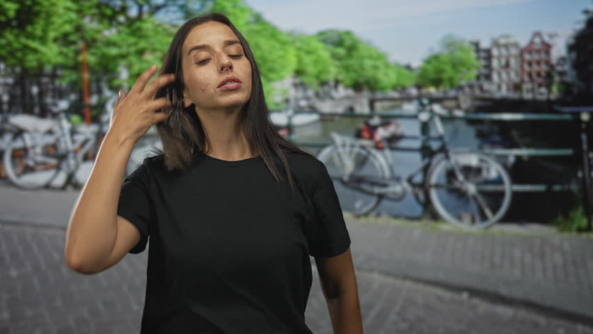 Woman standing by a parked bicycle on street in amsterdam, tilts head and looks aside; quiet contemplation.