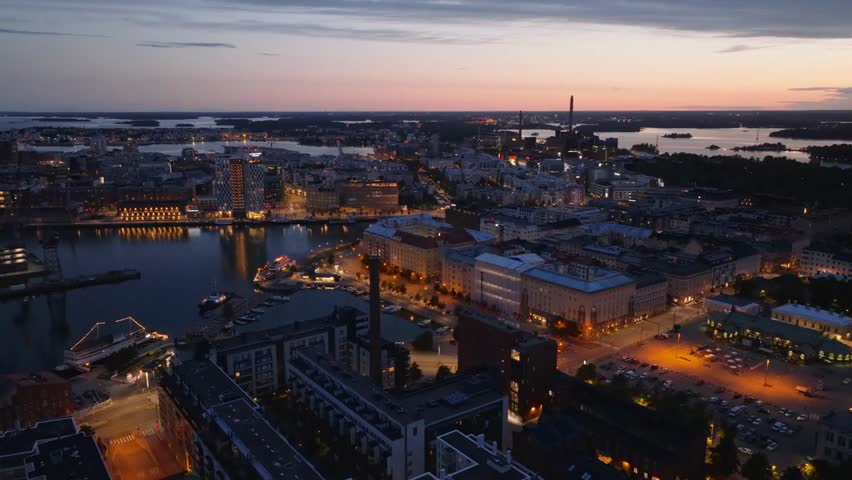 Forwards flyover of Helsinki at dusk, showing cars driving along a waterfront road with modern buildings in the city’s former port area.