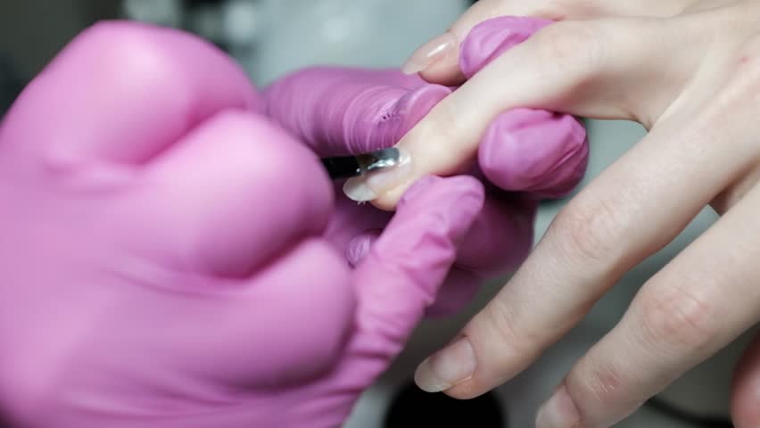 Professional close-up footage of a skilled nail technician wearing protective rubber gloves applying nail gel polish or base coat, making a manicure on a female client. Set in a bright, modern beauty salon, this slow-motion clip captures the wellness care and hygiene of luxury nail services. Ideal for content related to beauty industry standards, spa wellness, self-care routines, and professional cosmetic procedures.