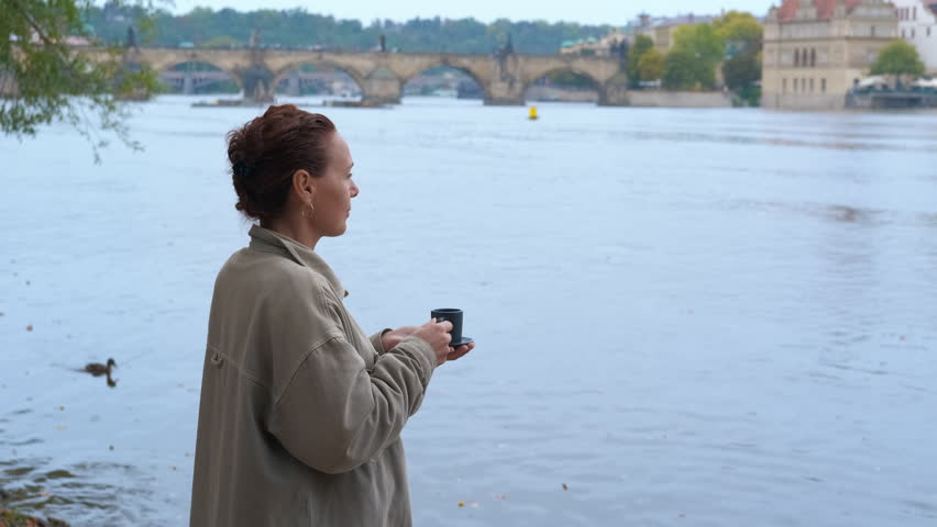 Woman enjoying coffee in Prague by the river. Female tourist savoring hot coffee while overlooking scenic vltava river, historic prague bridges, and architectural landmarks under cloudy autumn skies